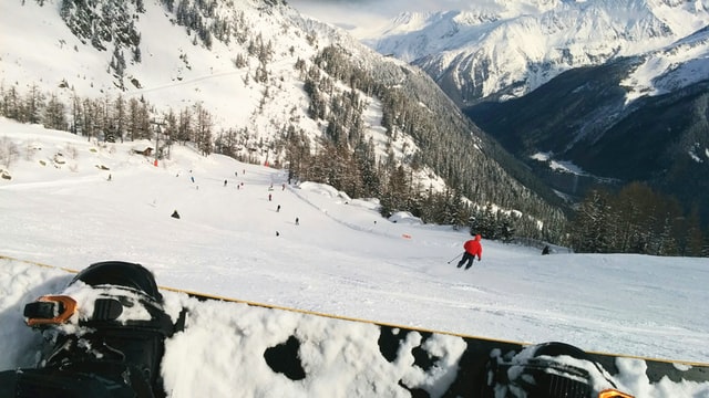 Sitting snowboarder watching a skier in a red jacket skiing down a piste