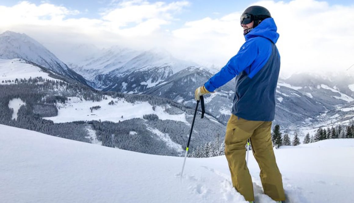 Male skier wearing a blue ski jacket standing at the top of a snowy hill looking down into a forest