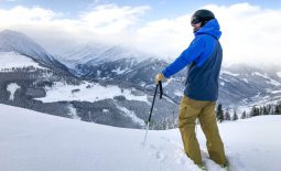 Male skier wearing a blue ski jacket standing at the top of a snowy hill looking down into a forest