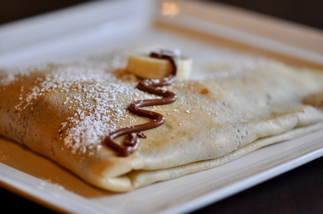 Close up of a crepe on a plate, sprinkled with icing sugar and chocolate sauce.