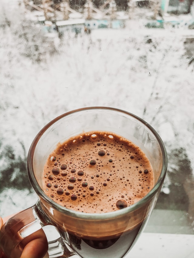 A hand holding a clear mug of hot chocolate against a snowy background