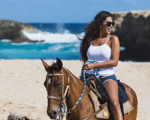Woman sat on a horse and smiling as she rides it along the beach 