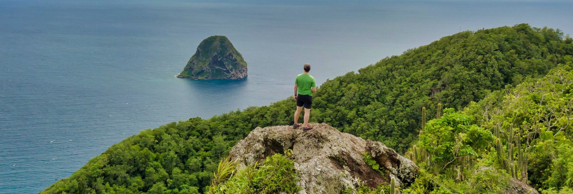 Man standing on top of a green tropical mountain whilst looking out to the sea