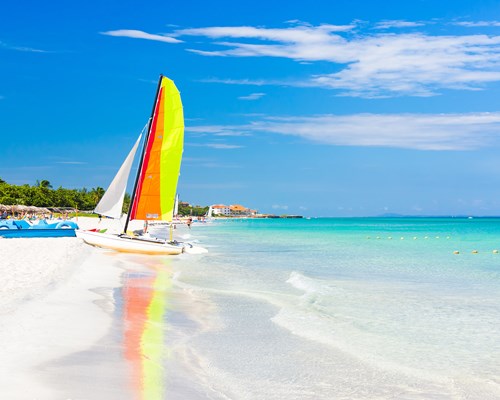 Sailing boat with colourful sail reflecting in the calm, clear blue sea next to a white sand beach