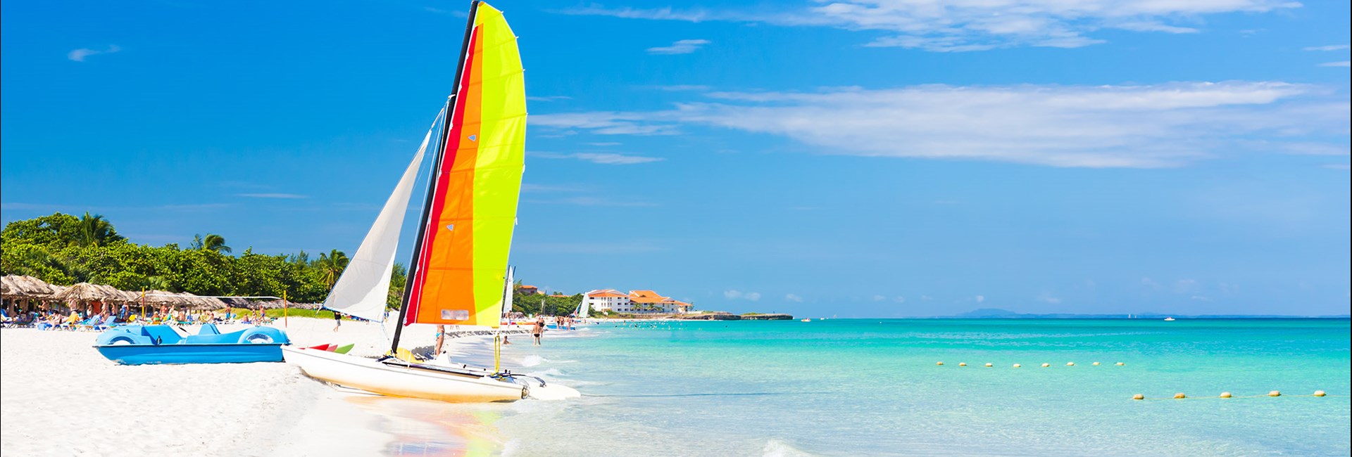  Sailing boat with colourful sail reflecting in the calm, clear blue sea next to a white sand beach 