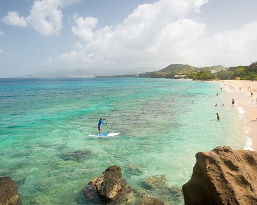 Man paddleboarding in clear tropical sea towards a white sand beach