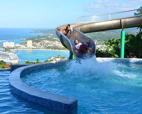 2 people coming down a large metal water slide with a view of a port in the background