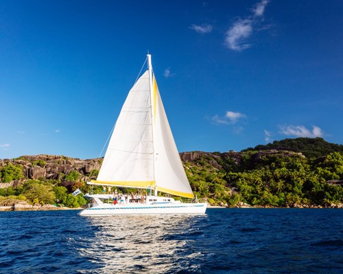White sailing boat with large white sails on blue ocean with leafy green hills in background - Praslin 