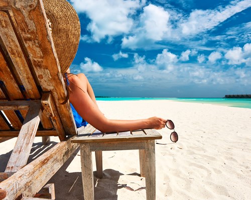 Woman holding a pair of sunglasses sitting on a wooden deck chair on a white sand beach 