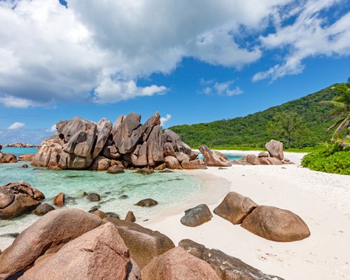 Granite boulders on white sand and crystal clear waters with hilly forest in background - Anse Cocos