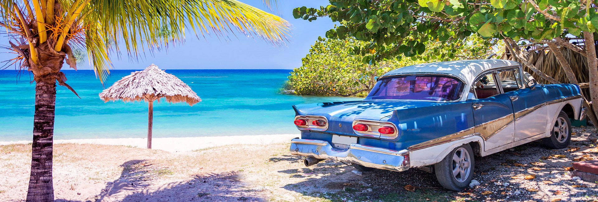 Vintage American blue car parked next to a palm tree on a white sand beach