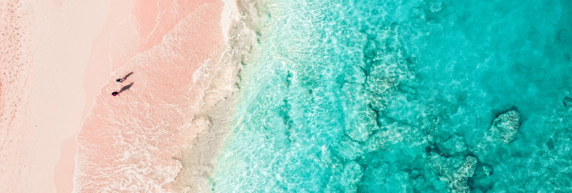 Aerial view of two people walking across a pink sand beach next to clear blue sea