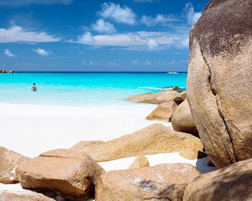 White sandy beach with lone man swimming in blue sea with rocks in foreground - Anse Georgette