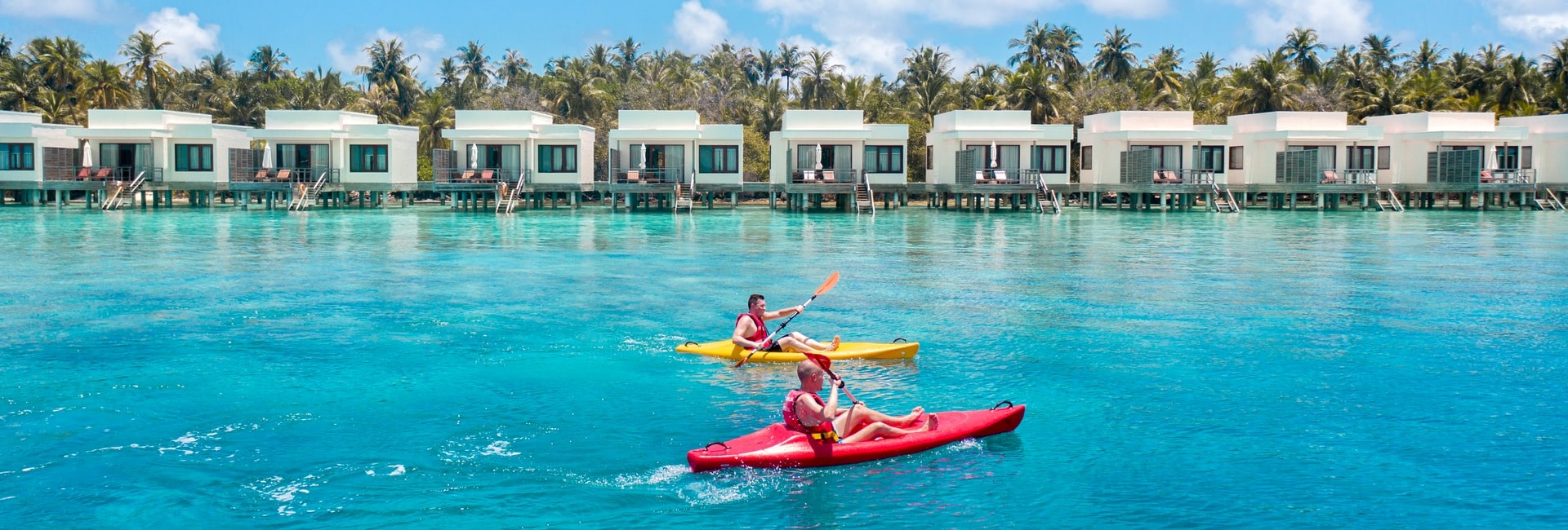 Two men in kayaks on bright blue sea going past overwater villas on a small island