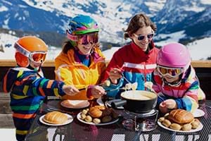A family with young kids, wearing brightly coloured ski jackets, sitting outdoors on a terrace eating cheese fondue